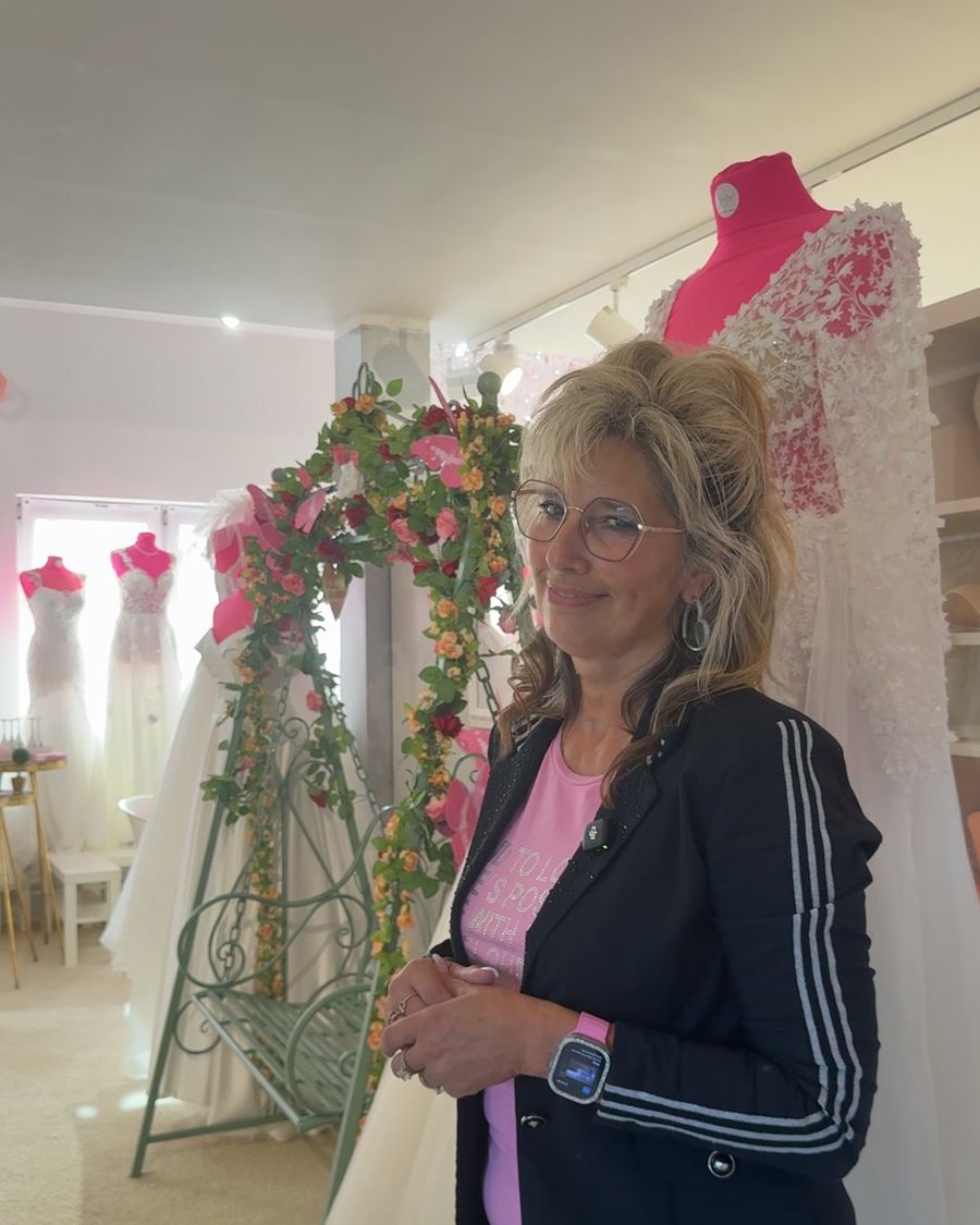 Yvonne im Laden vor dem Blumenbogen, neben einem Brautkleid mit Blütenapplikationen